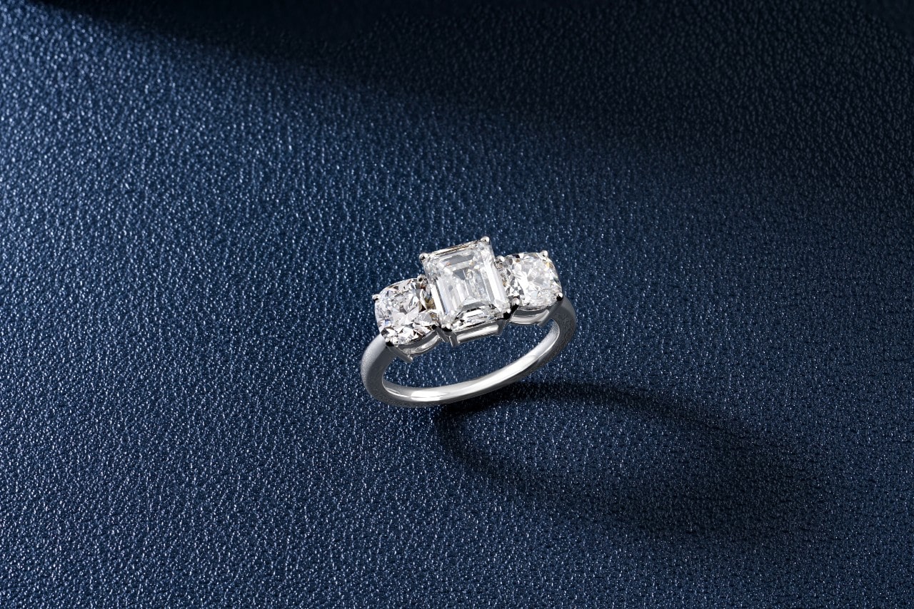A three-stone engagement ring with an emerald cut diamond center stone on black leather.