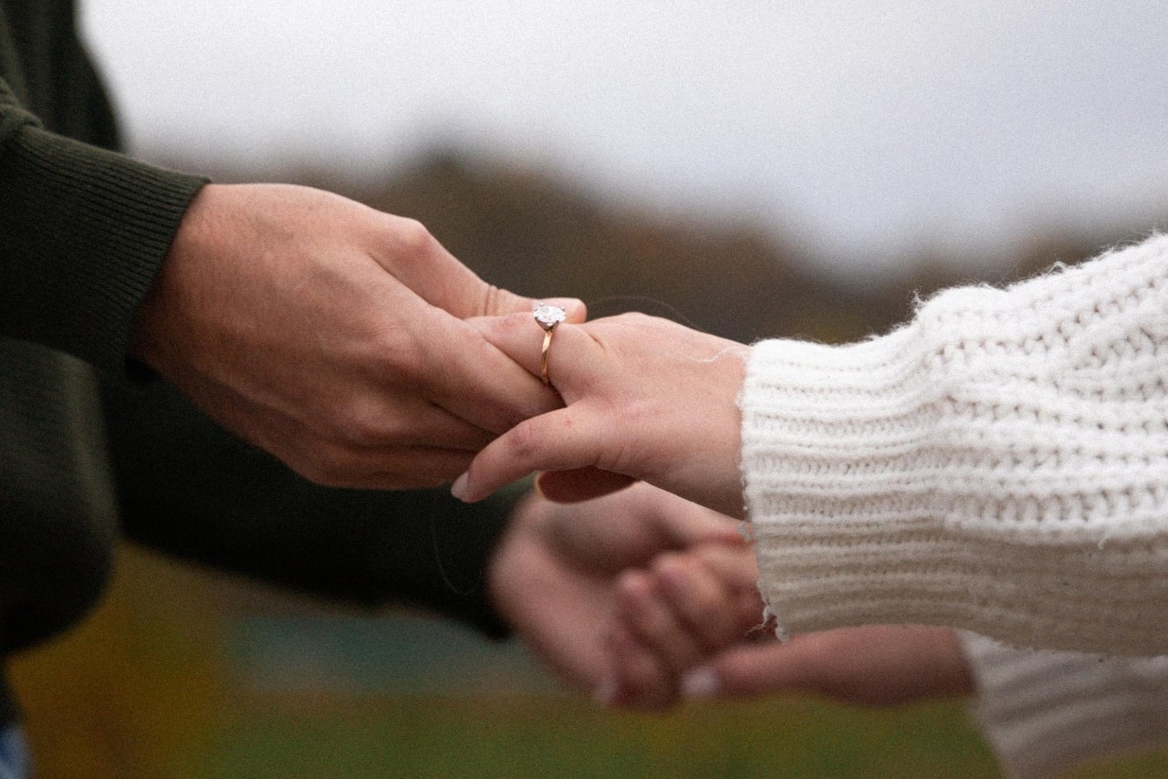 A couple holding hands in a foggy field, the woman wearing a yellow gold solitaire engagement ring.