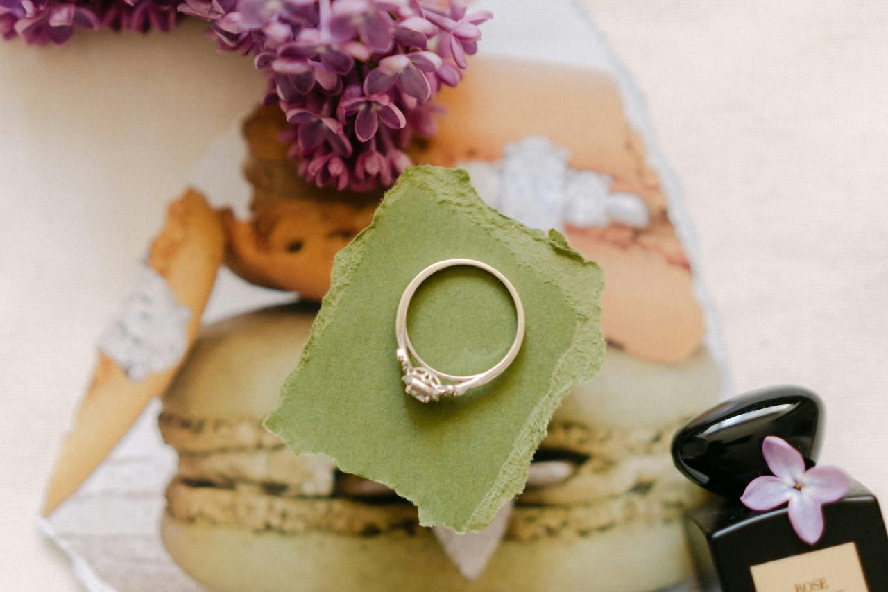 An engagement ring laying on top of a green square of paper, raised above a picture clipping of macaroons and some purple flowers surrounding.