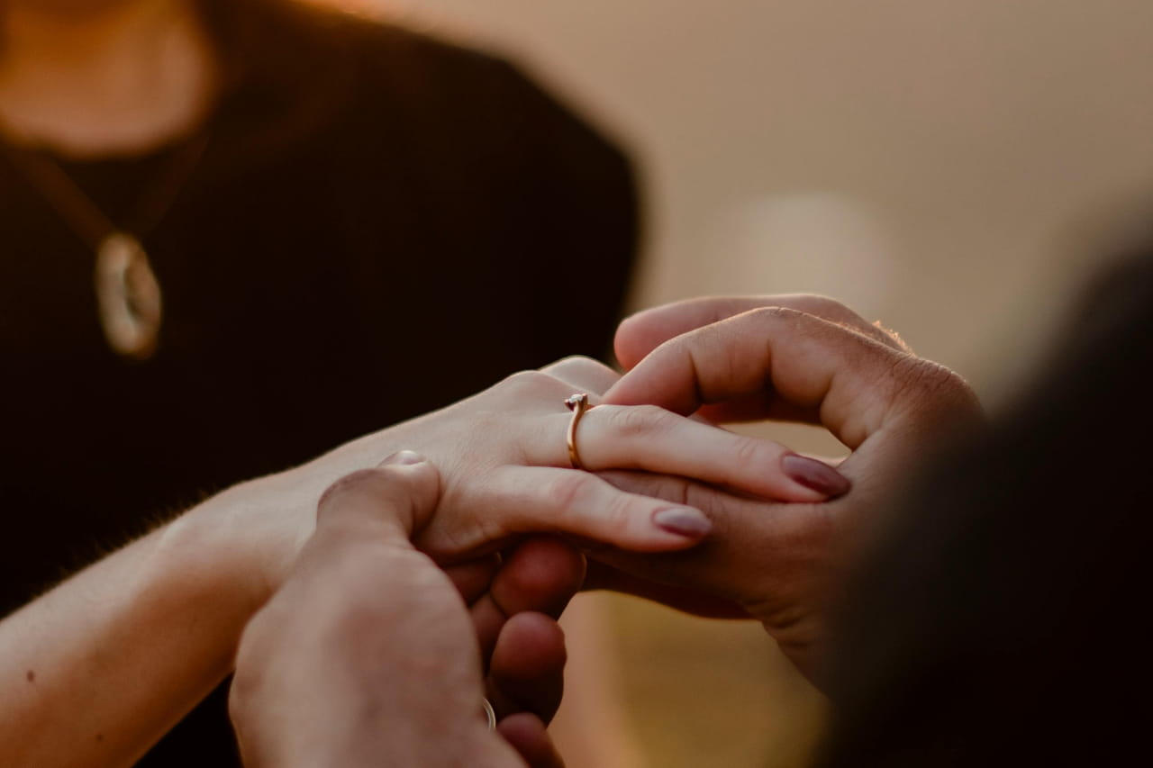 A man sliding a yellow gold engagement ring onto a woman’s finger, her silhouette blurred in the background.