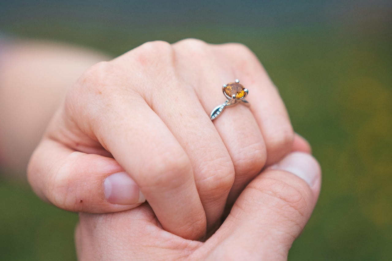 A close-up of a woman’s hand being held by another hand, showcasing her topaz-like engagement ring.