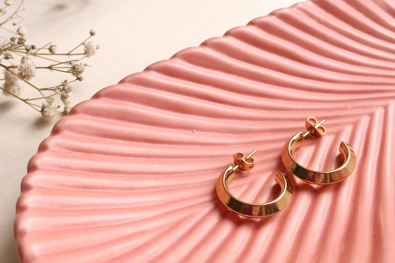 a pair of yellow gold huggie earrings lying on a pink shell dish