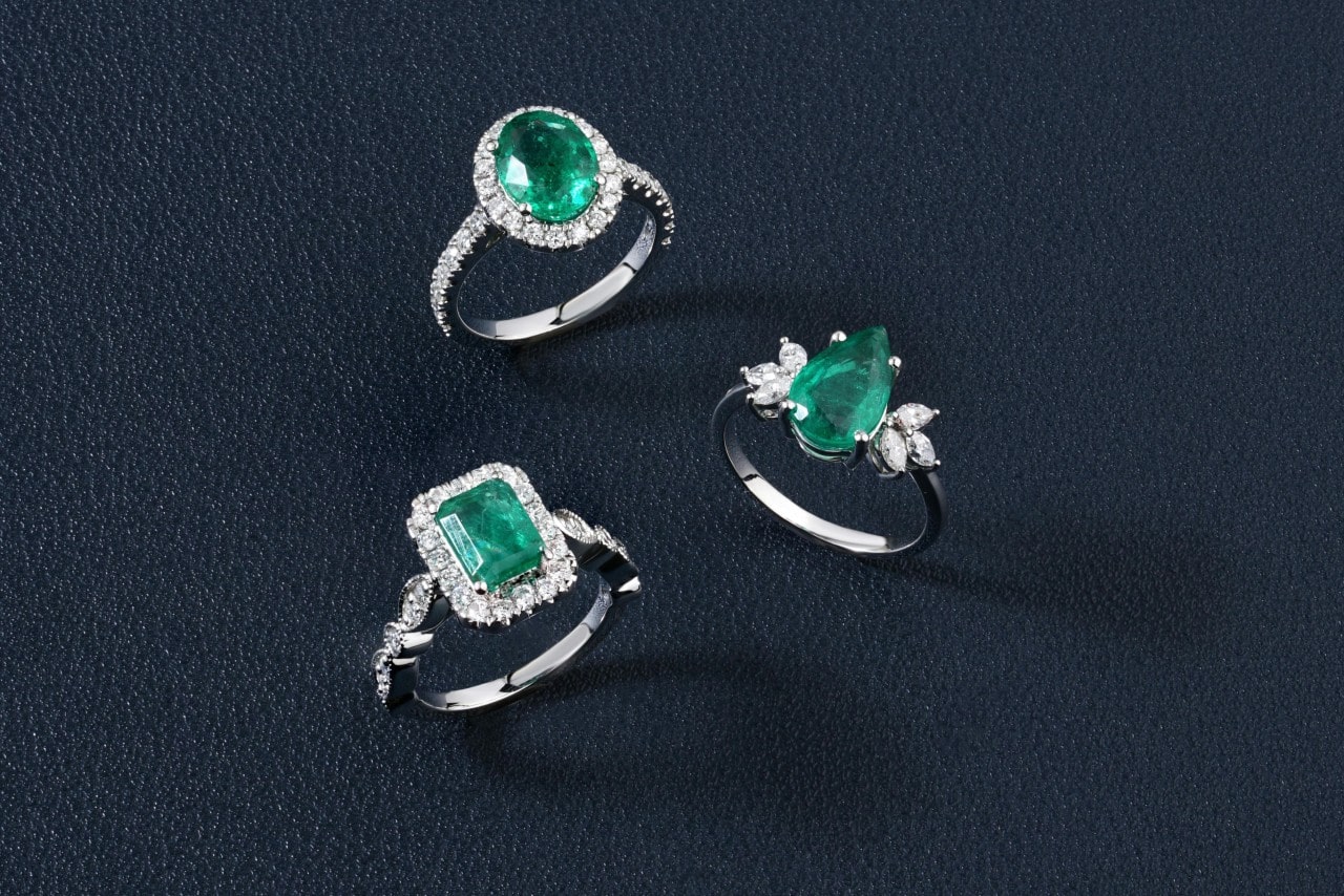 Three emerald engagement rings, a round cut, an emerald, and a pear, all sat on a leather surface.