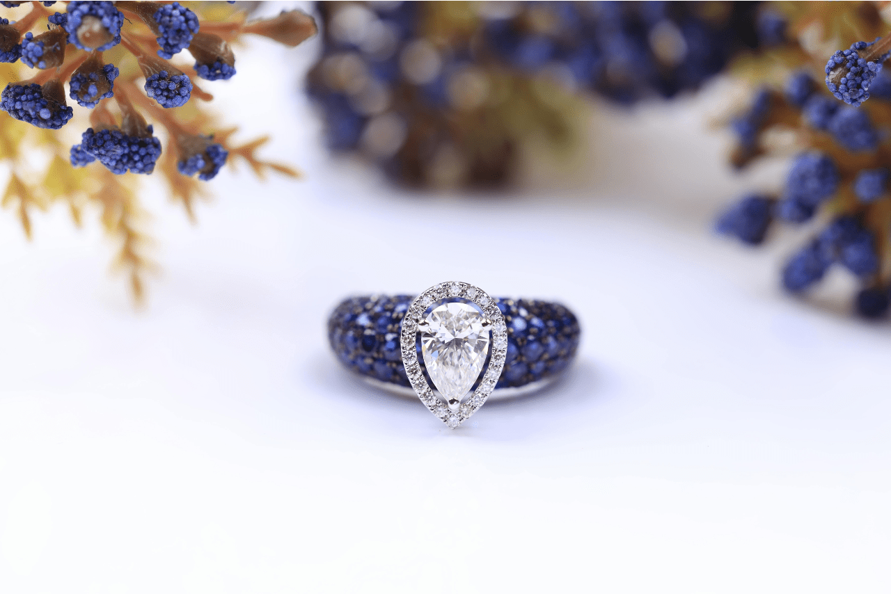 A beautiful pear-shaped engagement ring with a halo and thick band with dark blue diamonds sat on a white surface with some dark blue florals surrounding it.
