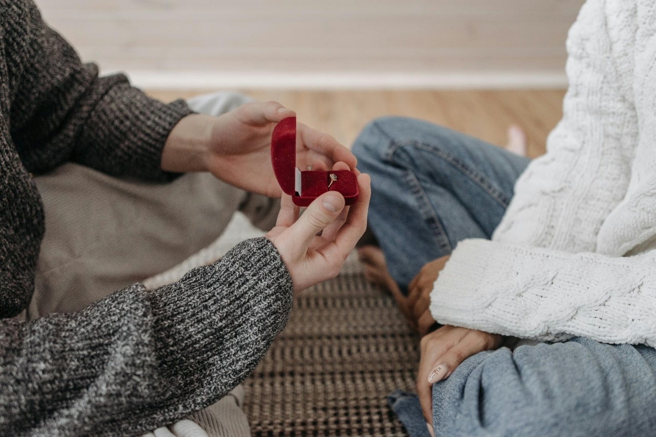 A close-up of a man proposing to his significant other in a casual setting, both wearing warm sweaters and sitting on a woven mat on a wooden floor.