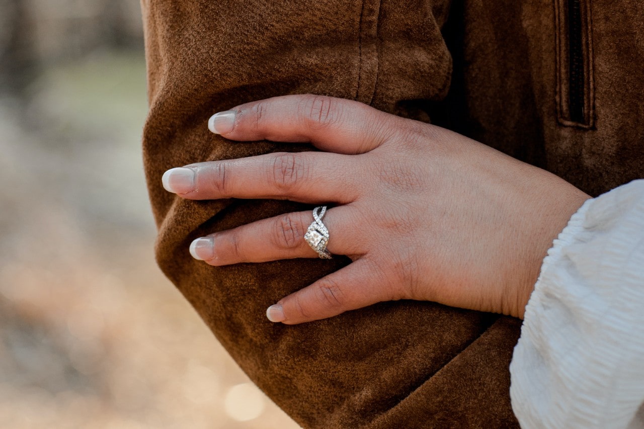 A close-up of a woman&rsquo;s hand adorned with an ornate halo engagement ring resting on her partner&rsquo;s arm in an outdoor area, both warmly dressed.