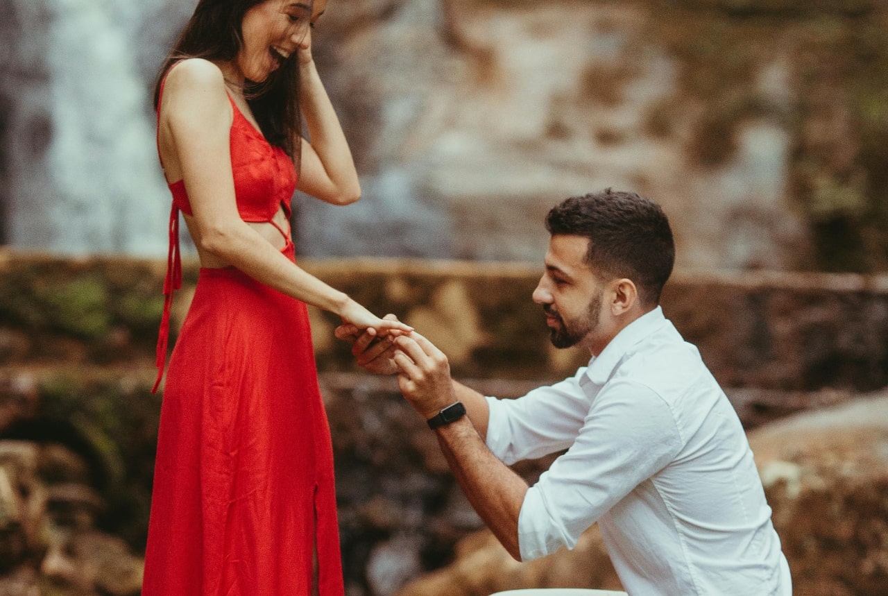 A view of a man proposing to his excited partner in a red dress, slipping the ring onto her finger.