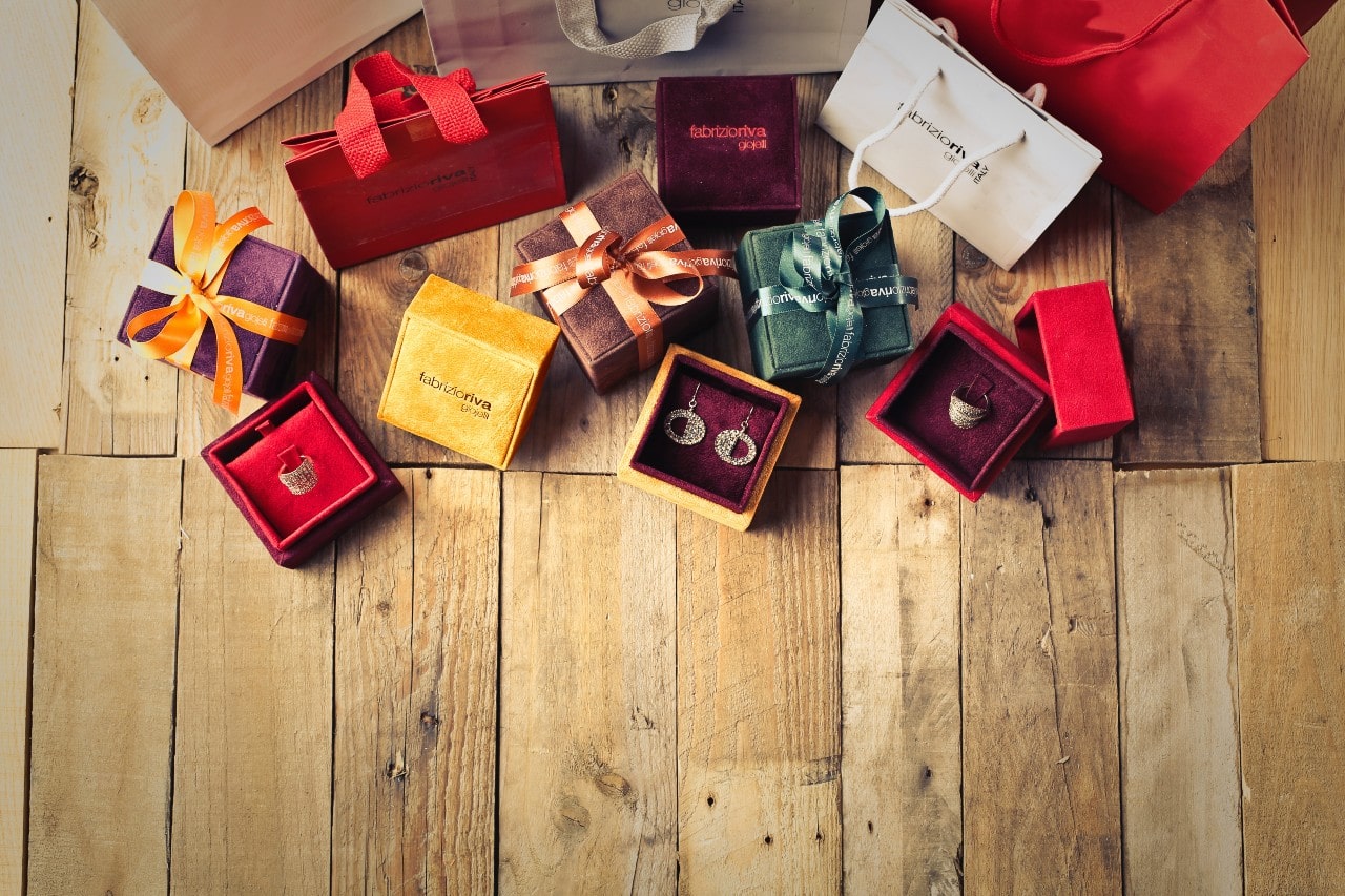 A colorful assortment of gift boxes and bags on an unfinished wooden floor, with some open boxes revealing elegant white gold jewelry.