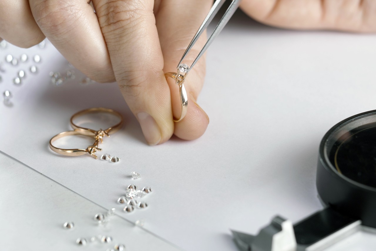 Close-up of a jeweler's hand uses tweezers to set a diamond on a gold ring.