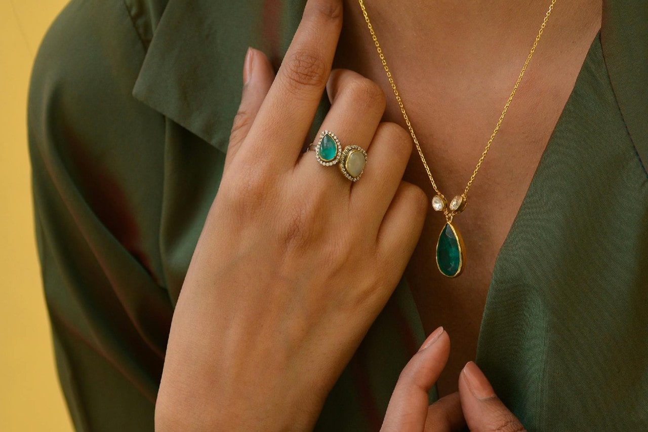Close-up of a person in a dark green blouse wearing a gold necklace with a green teardrop pendant and a two stone ring.