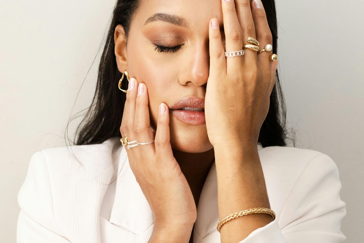 A woman in a white blazer covers one eye with her hand, adorned with fashion rings, earrings and bracelet.