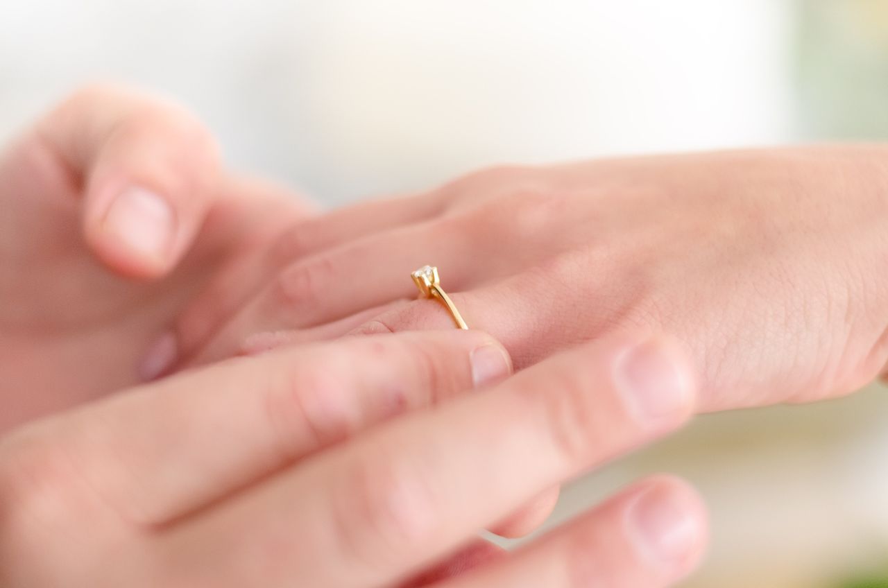 A hand is placing a yellow gold solitaire engagement ring with a single diamond onto another person's finger.