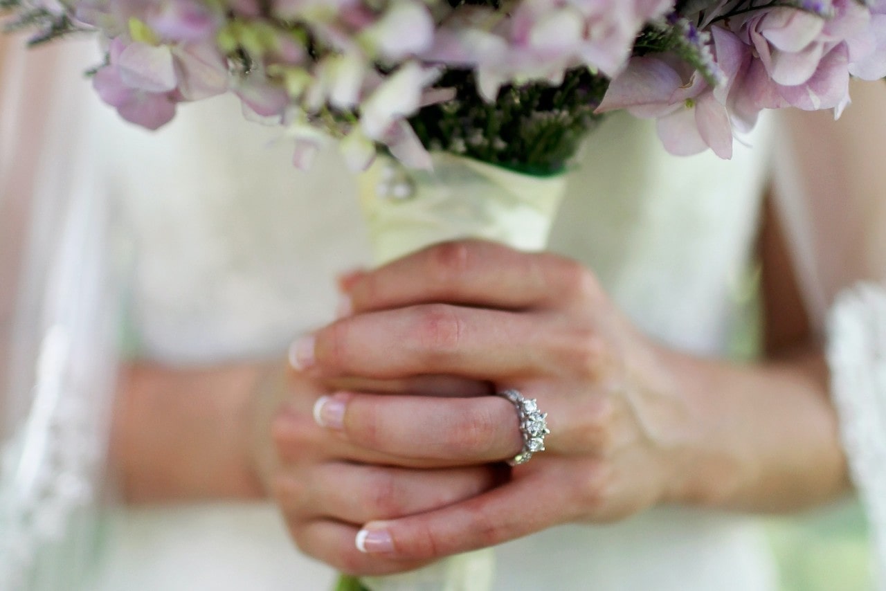 A close-up of a bride holding a bouquet of light purple flowers, showing her hands with a diamond engagement ring prominently displayed.