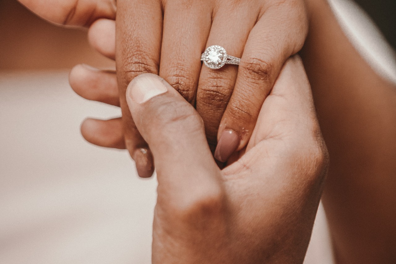 Close-up of a hand wearing a halo engagement ring with a round center diamond surrounded by smaller diamonds, held gently by another hand.