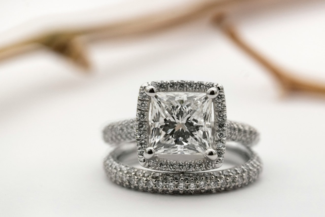 A close-up of a cushion-cut diamond side stone engagement ring with a matching diamond-studded wedding band, displayed on a white surface.