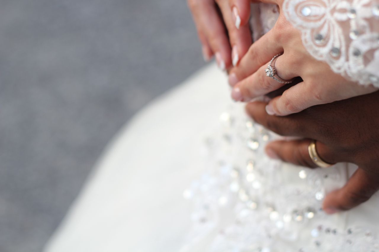 Close-up of a bride and groom's hands, with the bride's lace sleeve and engagement ring visible, gently clasping each other against a white wedding dress