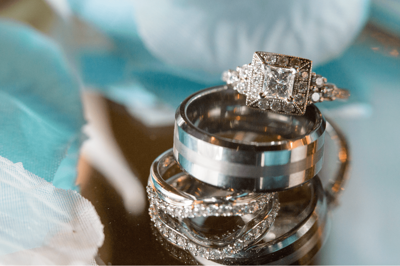 A stack of wedding rings on a reflective surface with a square-cut diamond ring sits atop two silver bands, surrounded by soft blue and white tulle.