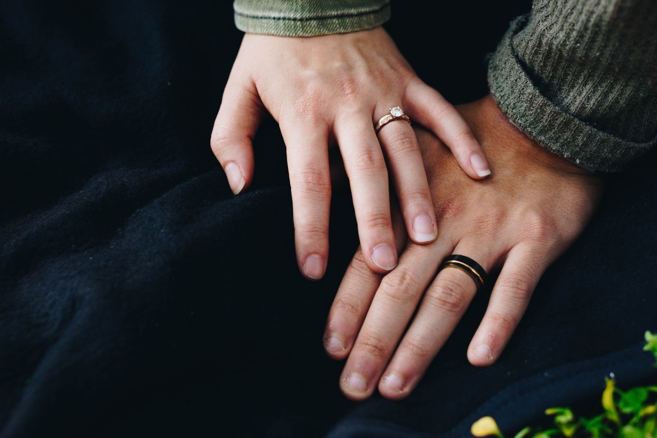 Close-up of two hands rest softly on dark fabric, one adorned with an engagement ring and the other with a wedding band, capturing a tender, intimate moment.