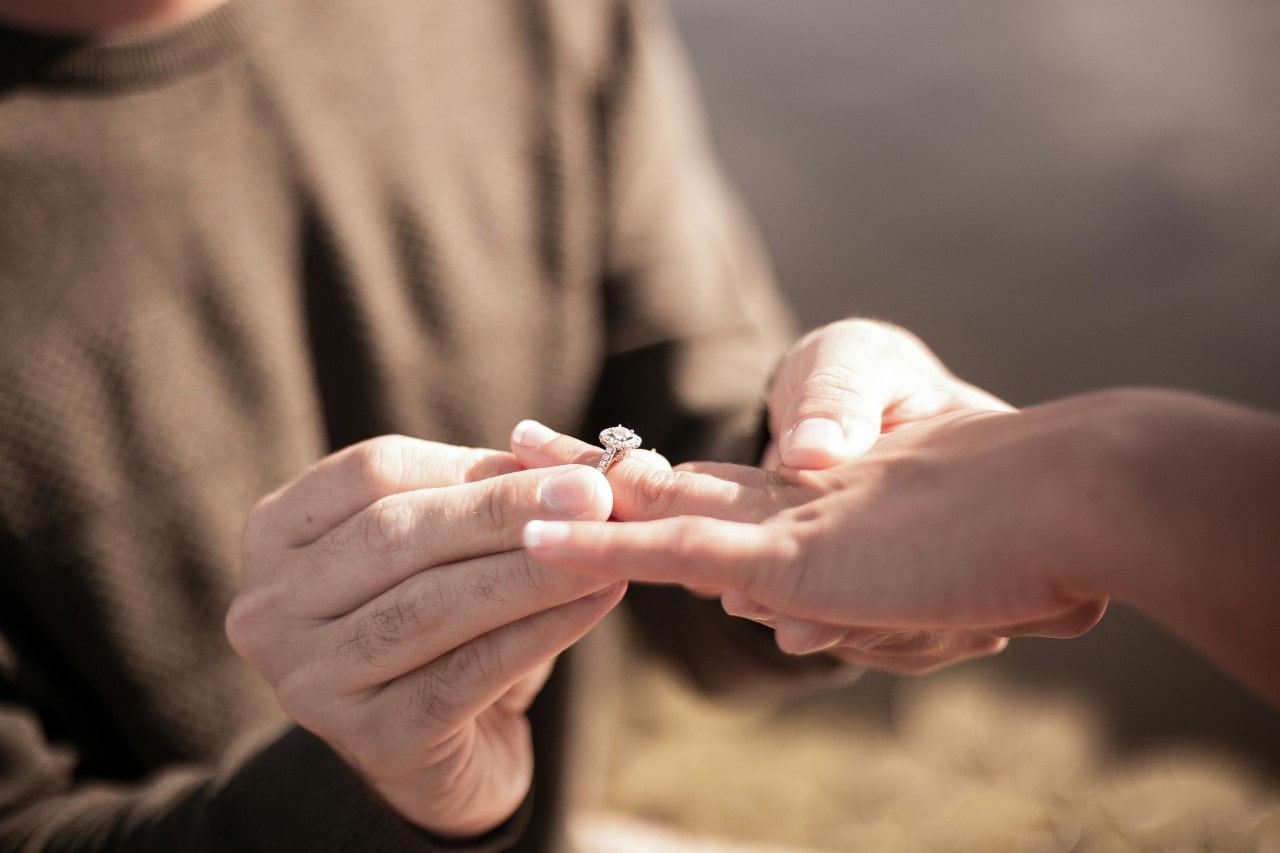 Close-up of a man in a sweater sliding a diamond engagement ring onto a woman's finger.