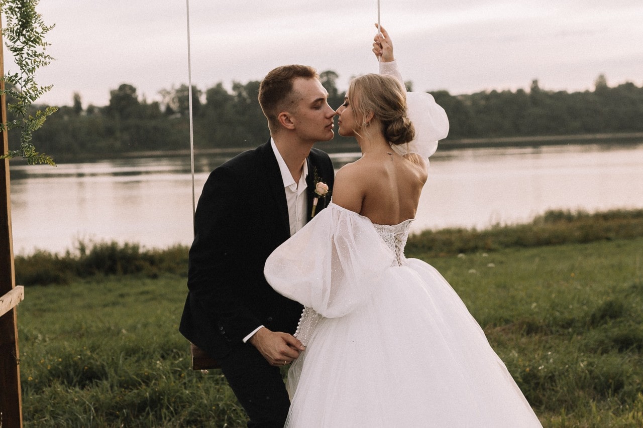 A bride and groom almost share a kiss while the groom sits on a swing, a lake view behind them.