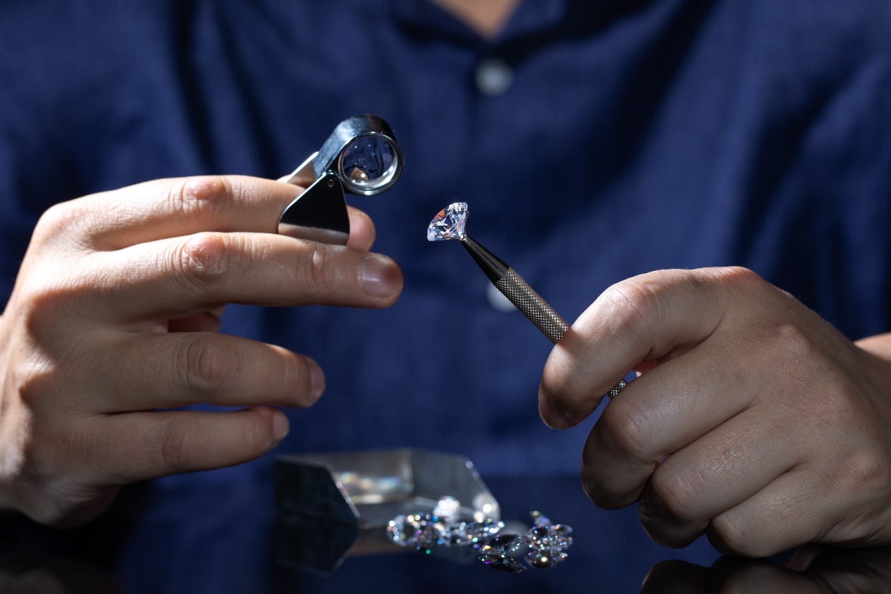 A close up of a jeweler in a blue shirt looking at a diamond through a magnifying loop.