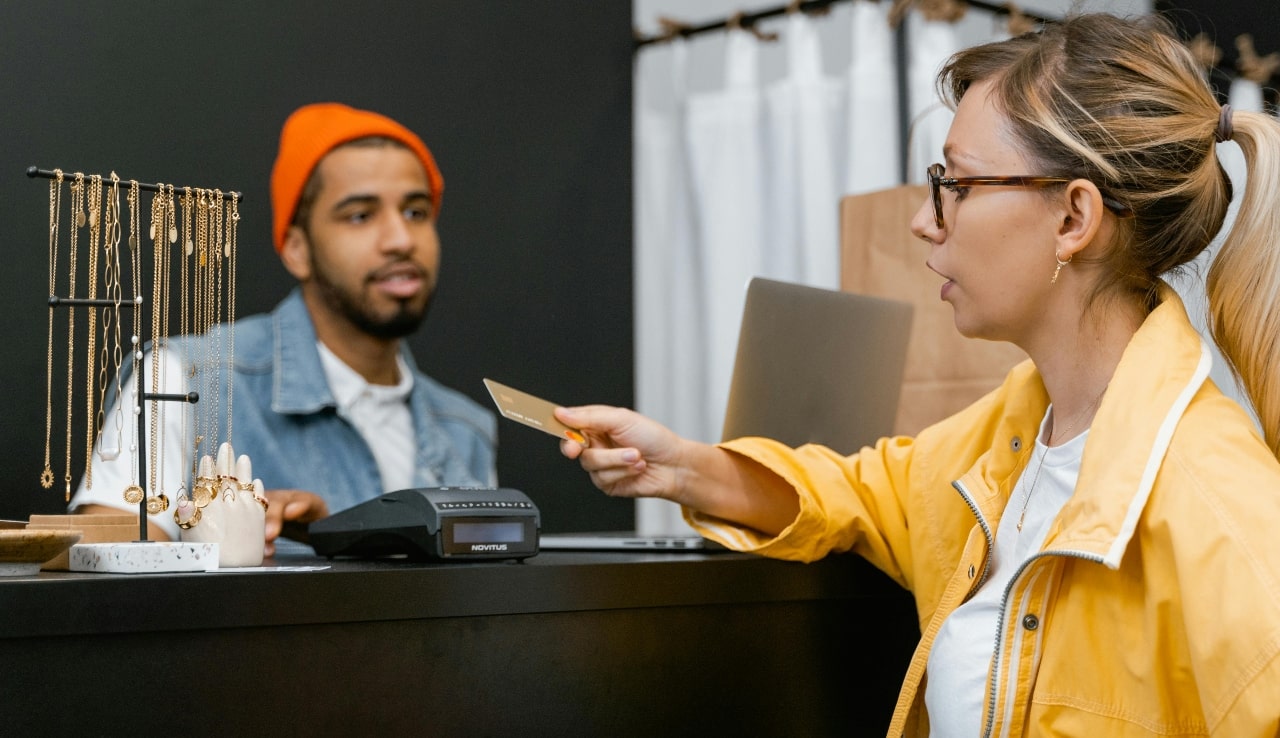 A woman in a yellow jacket pays with a credit card at a jewelry store counter while a cashier assists her.