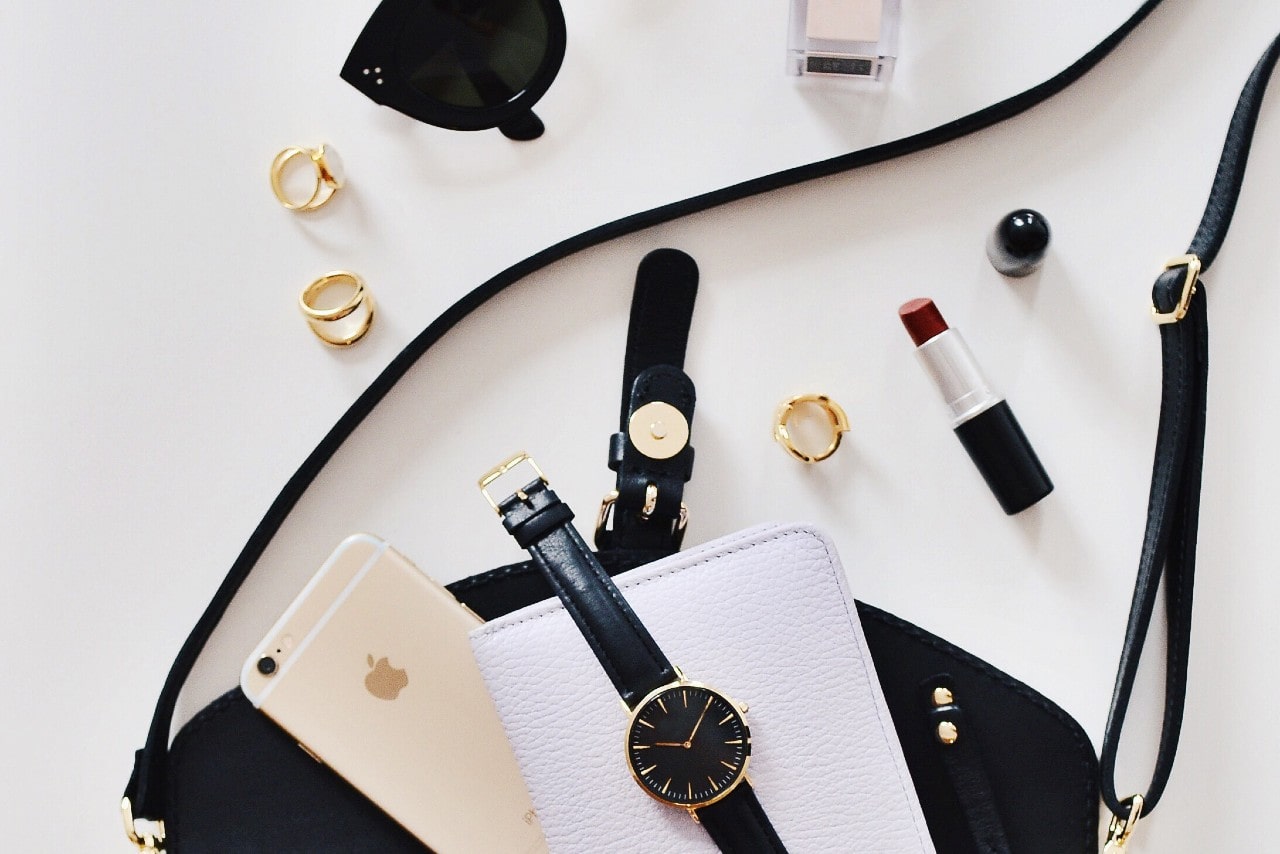 A purse sits on a table with a group of items including a watch, a phone, jewelry, and lipstick.