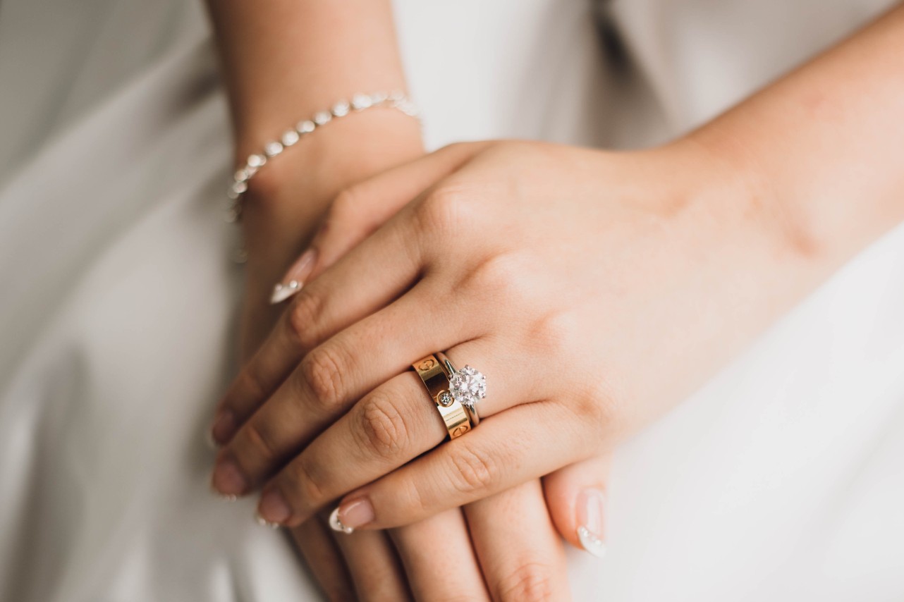 Close-up of bride’s hands with a gold wedding band and diamond solitaire engagement ring on one finger, manicured nails, wearing a delicate bracelet.