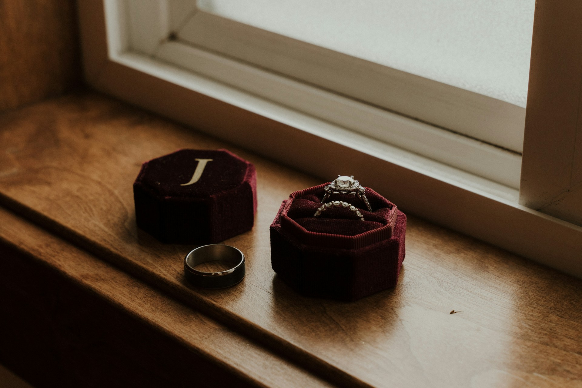 A silver wedding band and an ornate diamond engagement ring rest in a maroon velvet box with a 