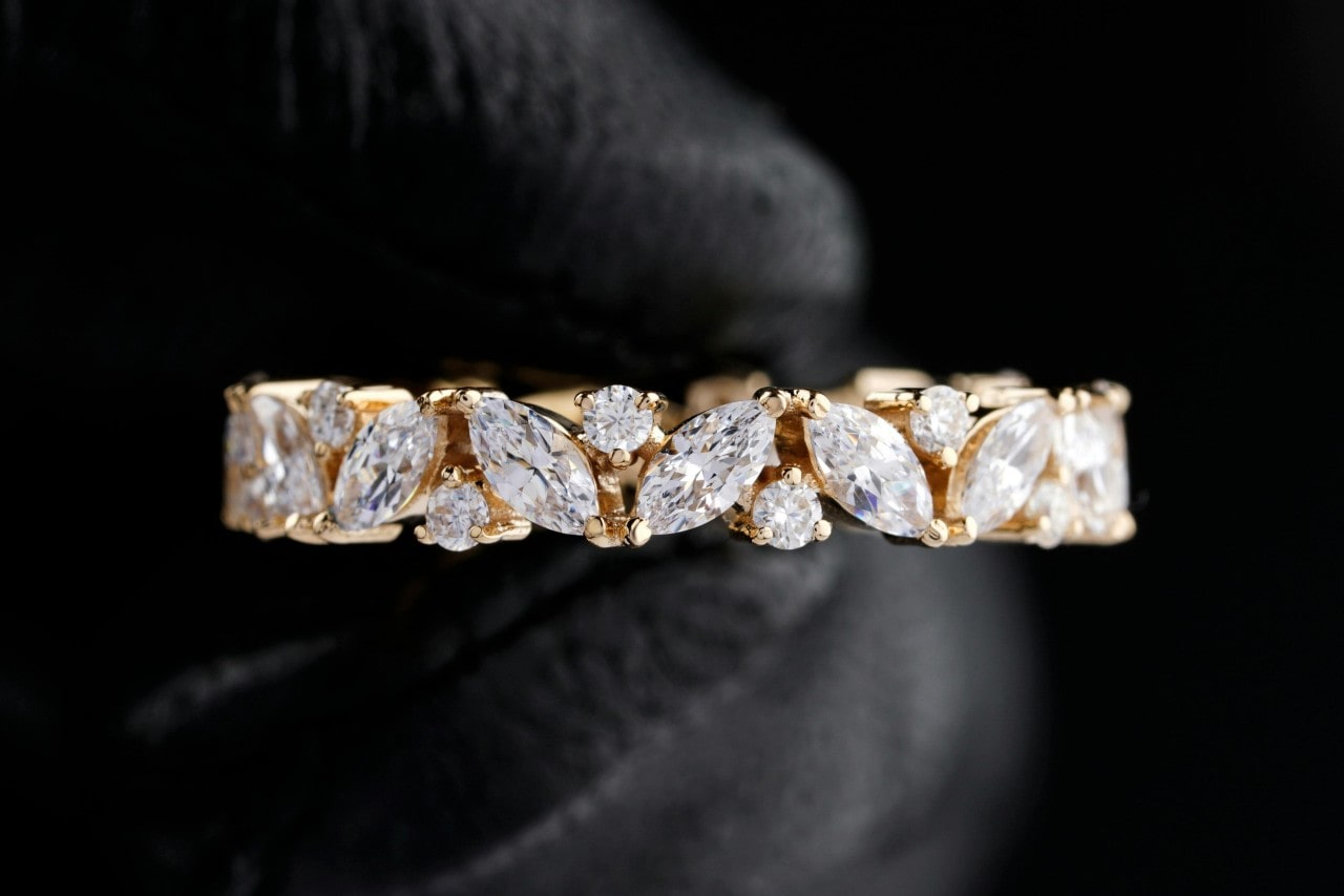A yellow gold ring adorned with alternating marquise and round-cut diamonds, held in a black gloved hand.
