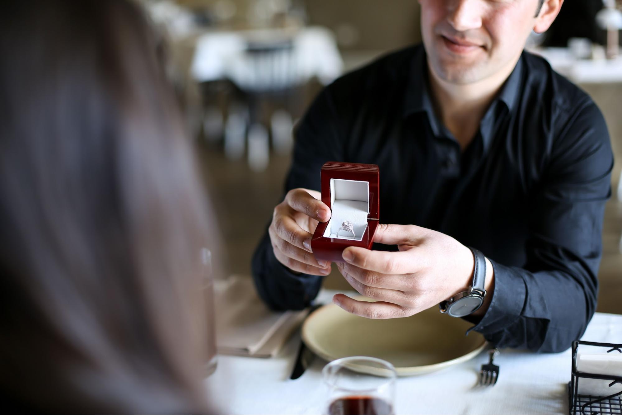 A couple sat at a table, the man holding open a red box with an engagement ring inside. A couple sat at a table, the man holding open a red box with an engagement ring inside.