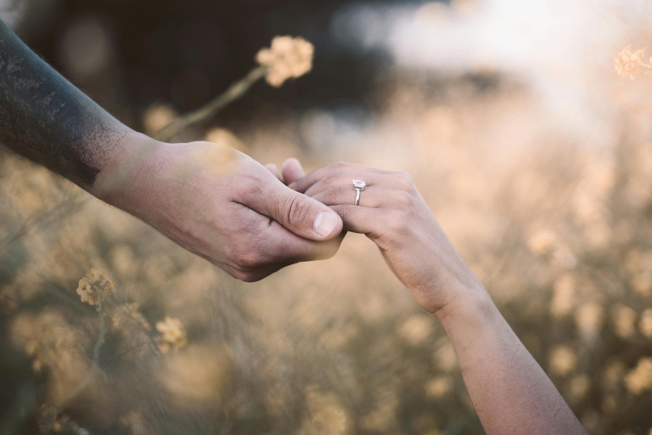 A couple holding hands in a field of yellow flowers, the woman showcasing her engagement ring. A couple holding hands in a field of yellow flowers, the woman showcasing her engagement ring.