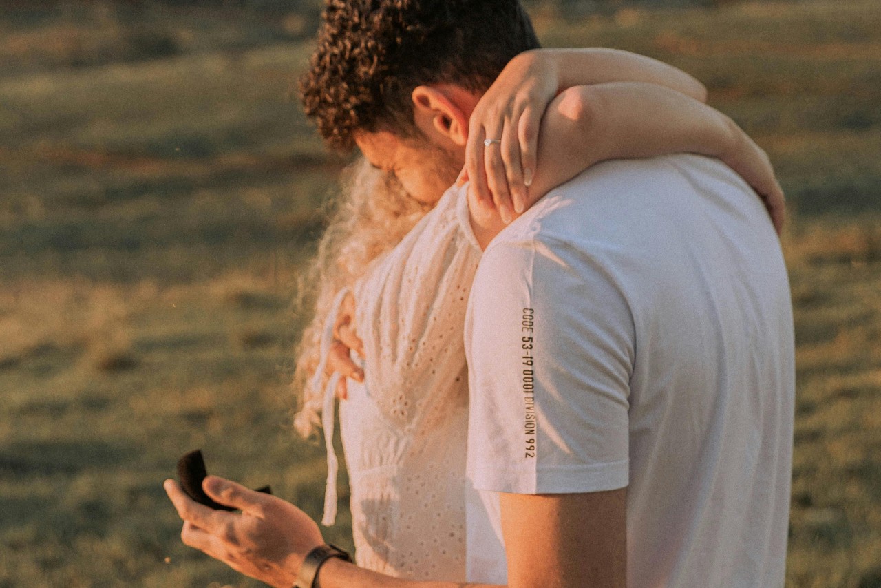 A couple embracing in an open field, the man holding open a ring box behind the woman’s back. A couple embracing in an open field, the man holding open a ring box behind the woman’s back.