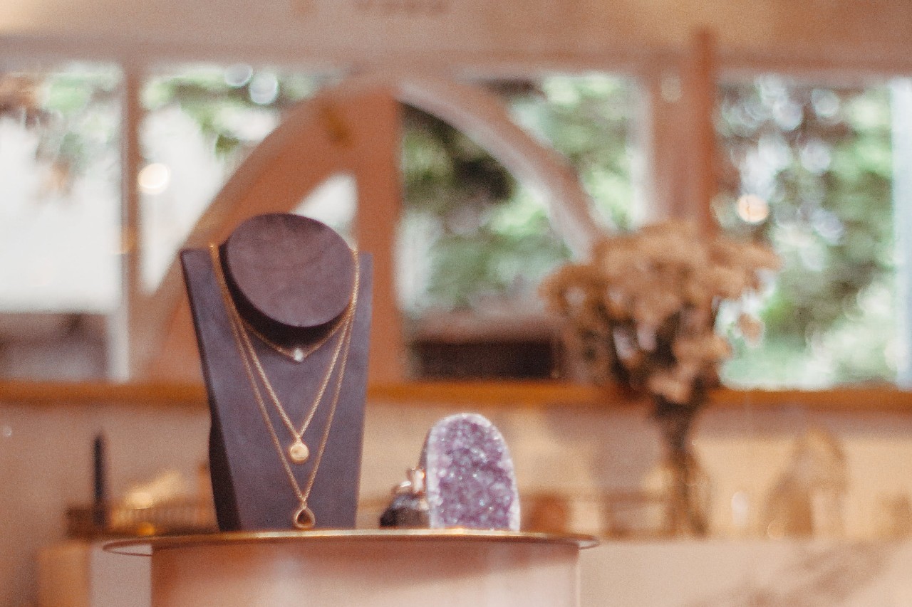 A close-up of a jewellery display with layered gold necklaces on a black stand inside a warmly lit boutique.
