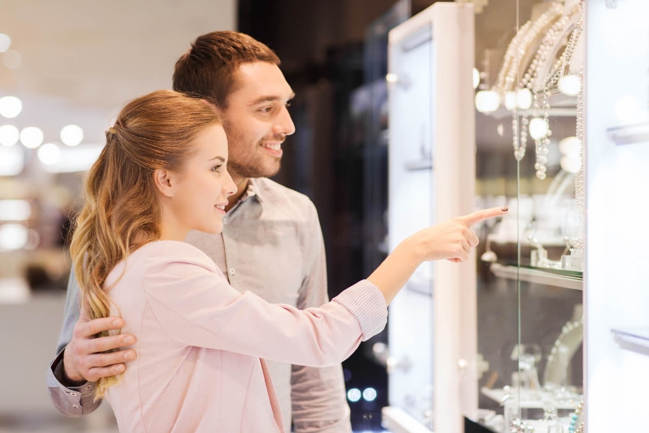 A couple standing in front of a jewellery shop window with a woman pointing at it, while a man smiling and embracing her.