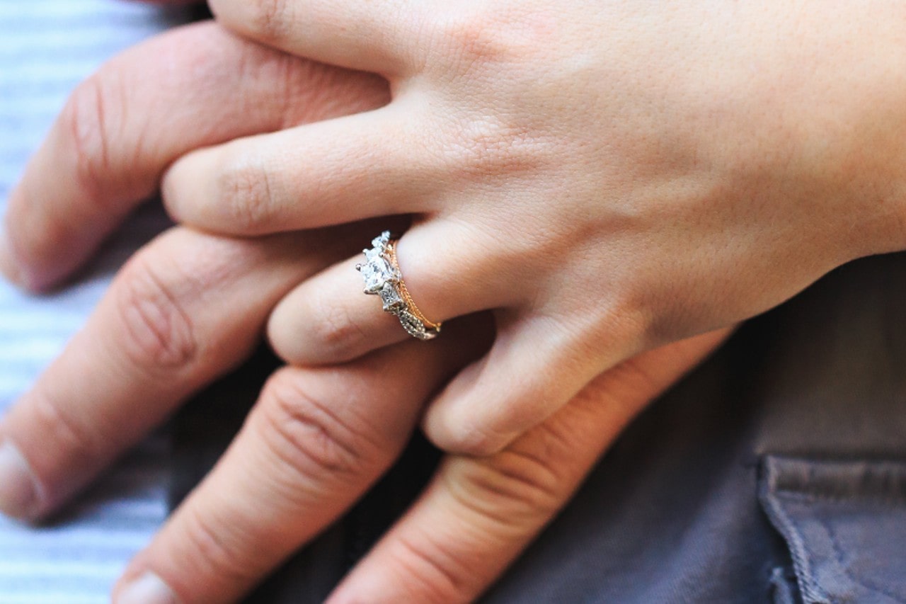 A close-up of a woman holding her boyfriend’s hand, with emphasis on the intricate engagement ring on her ring finger.