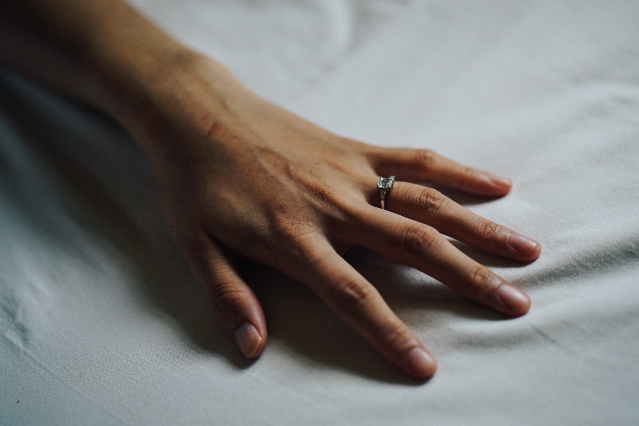 A close-up of a woman’s hand resting on a soft sheet, with emphasis on the engagement ring on her ring finger.