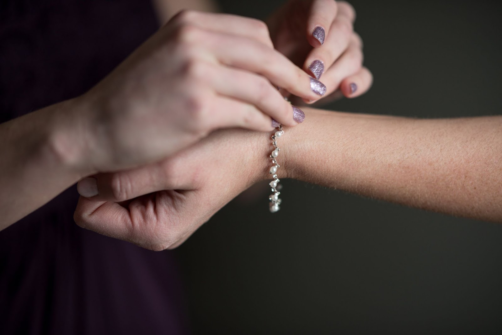 A bridesmaid wearing a plum formal dress fastens a latch on a pearl bracelet for the bride