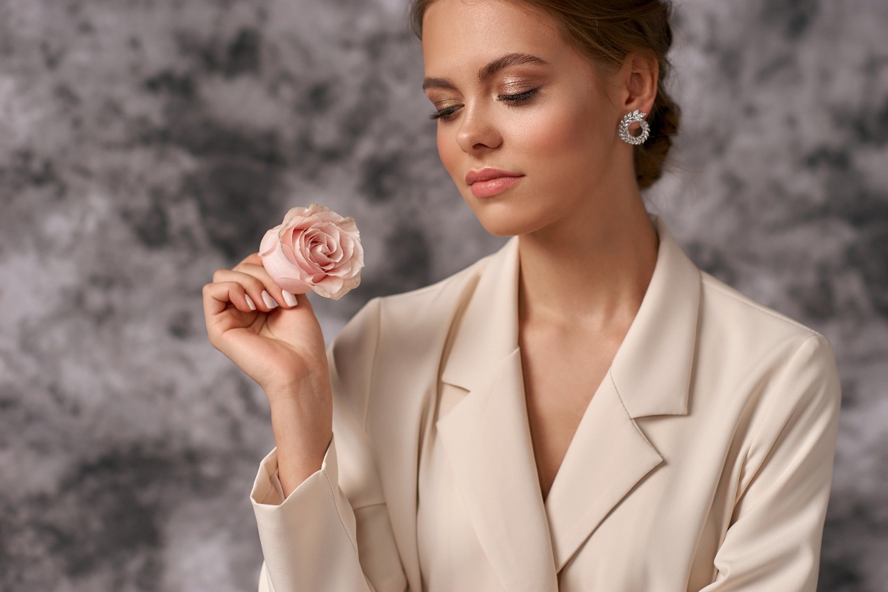 A woman in a beige blazer wearing diamond round earrings, holds a pink rose.