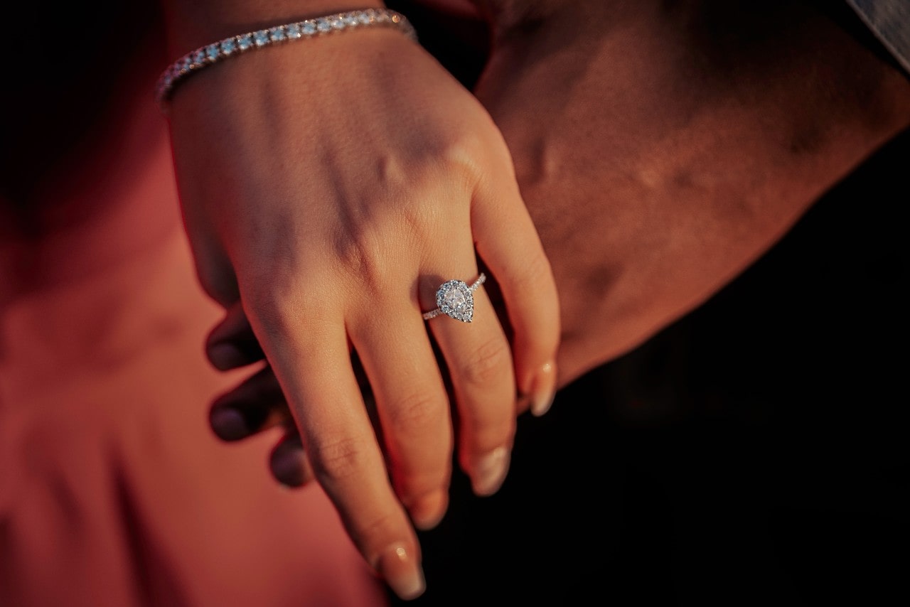 A close-up of the bride's hand with long nails, diamond tennis bracelet, and halo pear shaped engagement ring on top of the man’s hand against a blurry background of her pink dress.