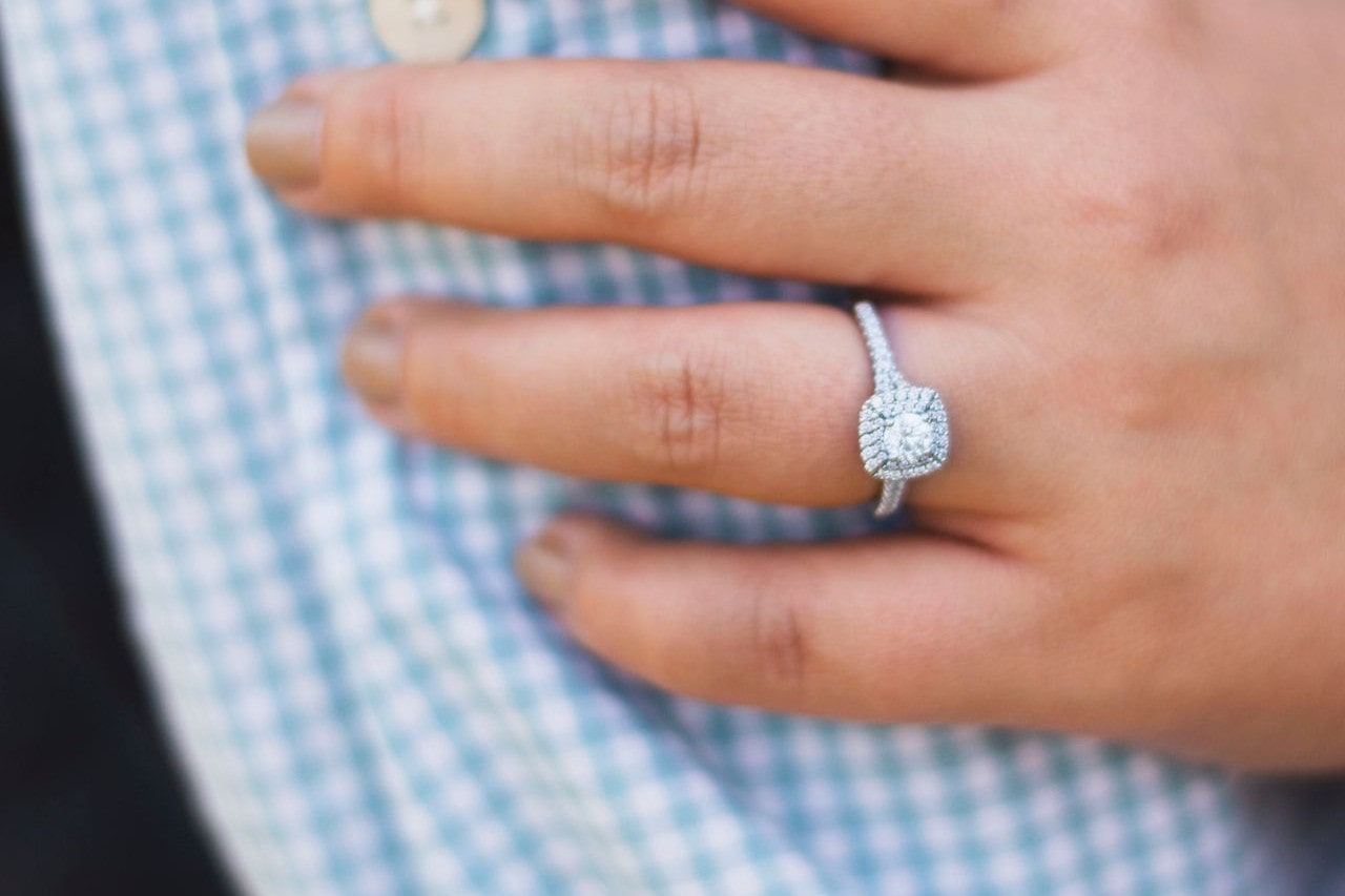 A close-up of a woman's hand wearing a sparkling cushion-cut diamond ring with a double halo design against a blue checkered shirt.