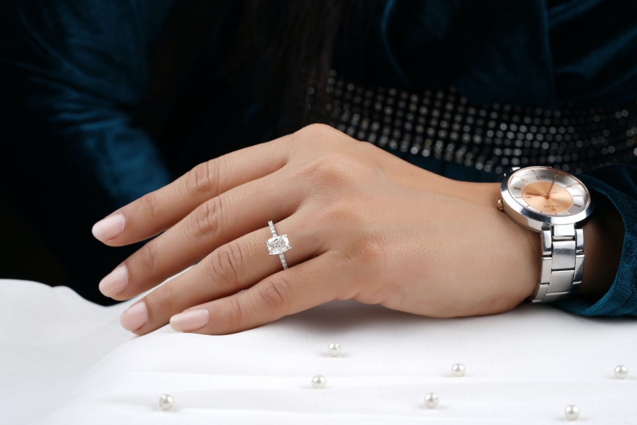 A woman's hand adorned with a rectangular-cut diamond ring and a silver wristwatch, resting on a white surface scattered with small pearls.